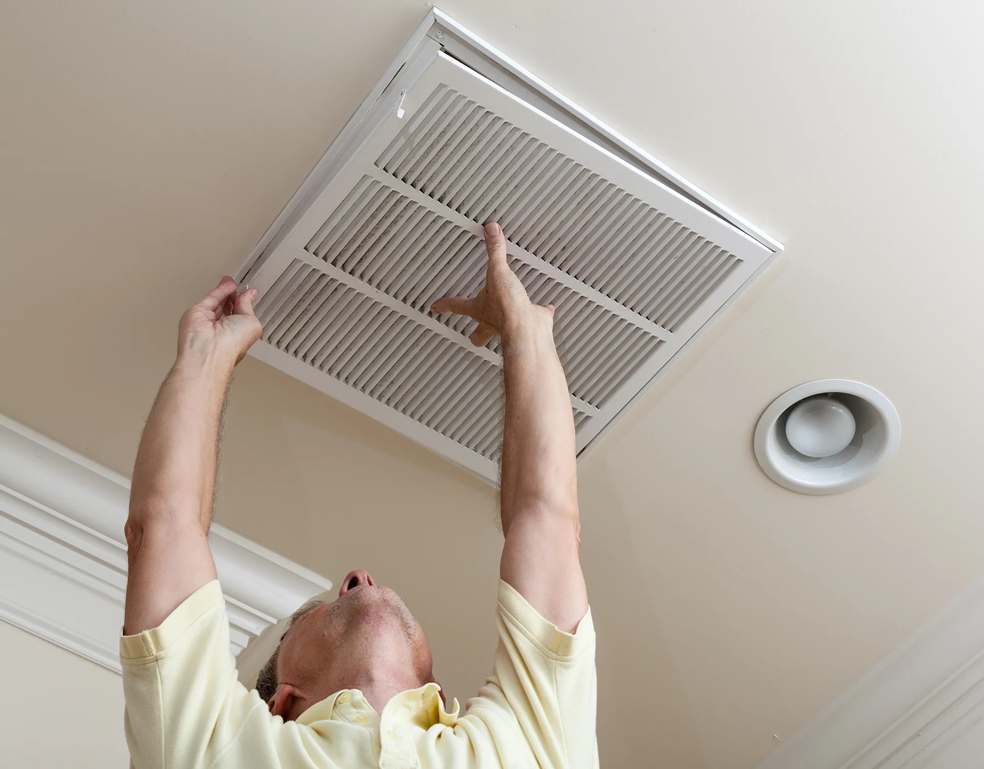 Up-close view of a person reaching up to open a ceiling-mounted air return vent to replace a dirty HVAC filter in a residential home.